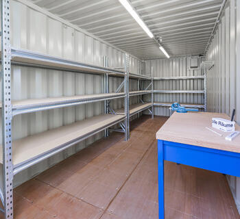 Åben lagercontainer med hylder og arbejdsbænk indvendigt.